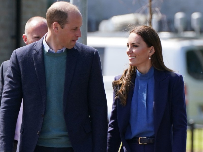 Prince William (L) wearing teal sweater and talking to Kate Middleton, who is wearing a blue top and blazer