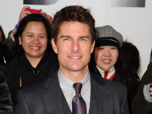 Tom Cruise smiles in a classic black suit and tie, with two women smiling behind him