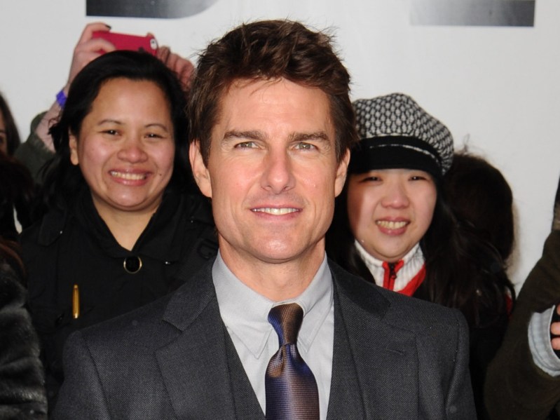 Tom Cruise smiles in a classic black suit and tie, with two women smiling behind him