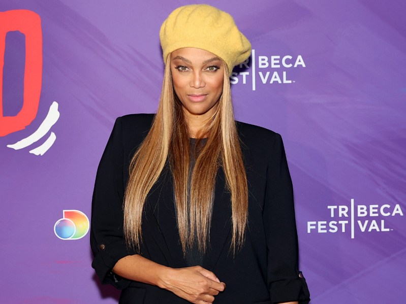 Tyra Banks smiles in black outfit with yellow hat against purple backdrop