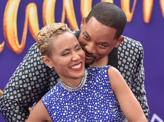 Jada Pinkett smiles with Will Smith behind her, his hands wrapped around her waist. He appears to be whispering something in her ear. Smith is wearing a patterned black and white blazer and Pinkett is wearing a purple dress