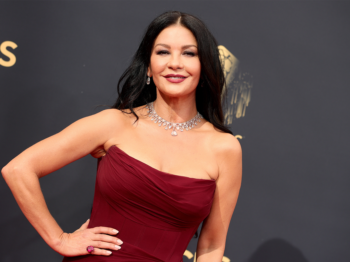 Catherine Zeta-Jones smiles with a hand on her hip in a strapless, dark red gown.