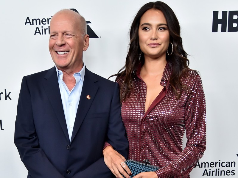 Bruce Willis (L) in navy suit jacket linking arms with his wife, Emma Heming, who is wearing a burgundy top