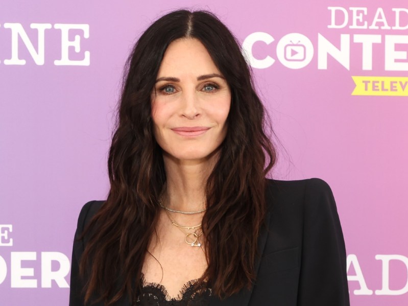 Courteney Cox smiling in black outfit against blue backdrop