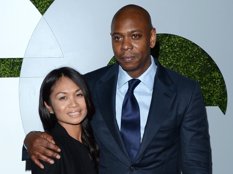Dave Chappelle (R) in navy blue suit standing with Elaine Chappelle who is viewed from the shoulders up against white backdrop
