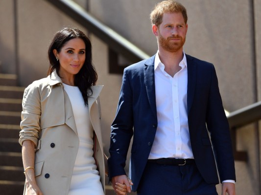 Meghan Markle (L) in white dress with tan coat walking next to Prince Harry, who is wearing a navy blazer over a white dress shirt