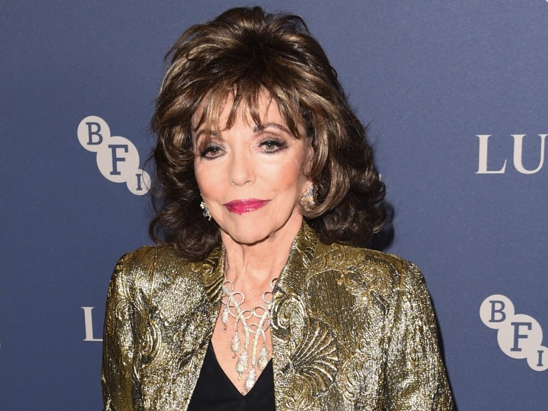 Joan Collins smiles in black shirt with gold jacket against gray-purple backdrop