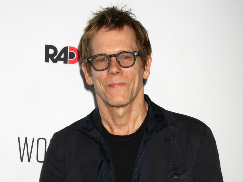 Kevin Bacon smirking in all-black outfit and glasses