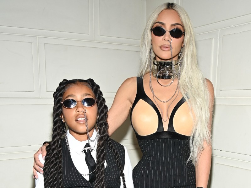North West, wearing a black pinstriped vest over a white shirt, stands with mother Kim Kardashian, in a black and flesh colored dress