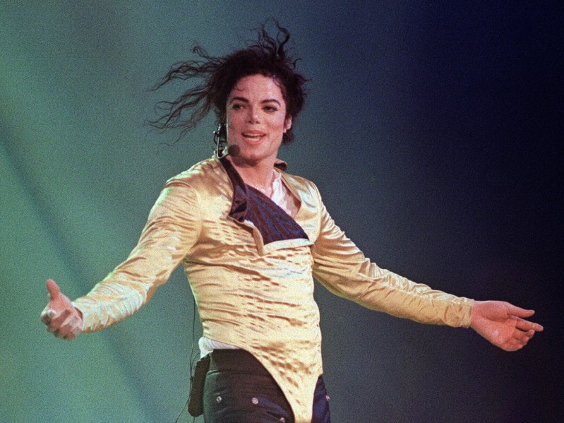 Michael Jackson standing onstage in gold jumpsuit against deep teal backdrop