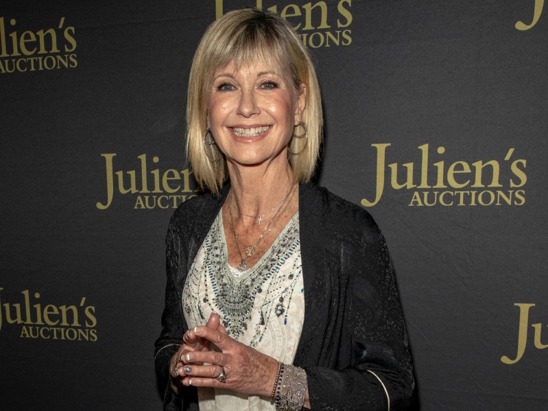 Olivia Newton-John smiles in white top with black cardigan. She is standing against a black backdrop