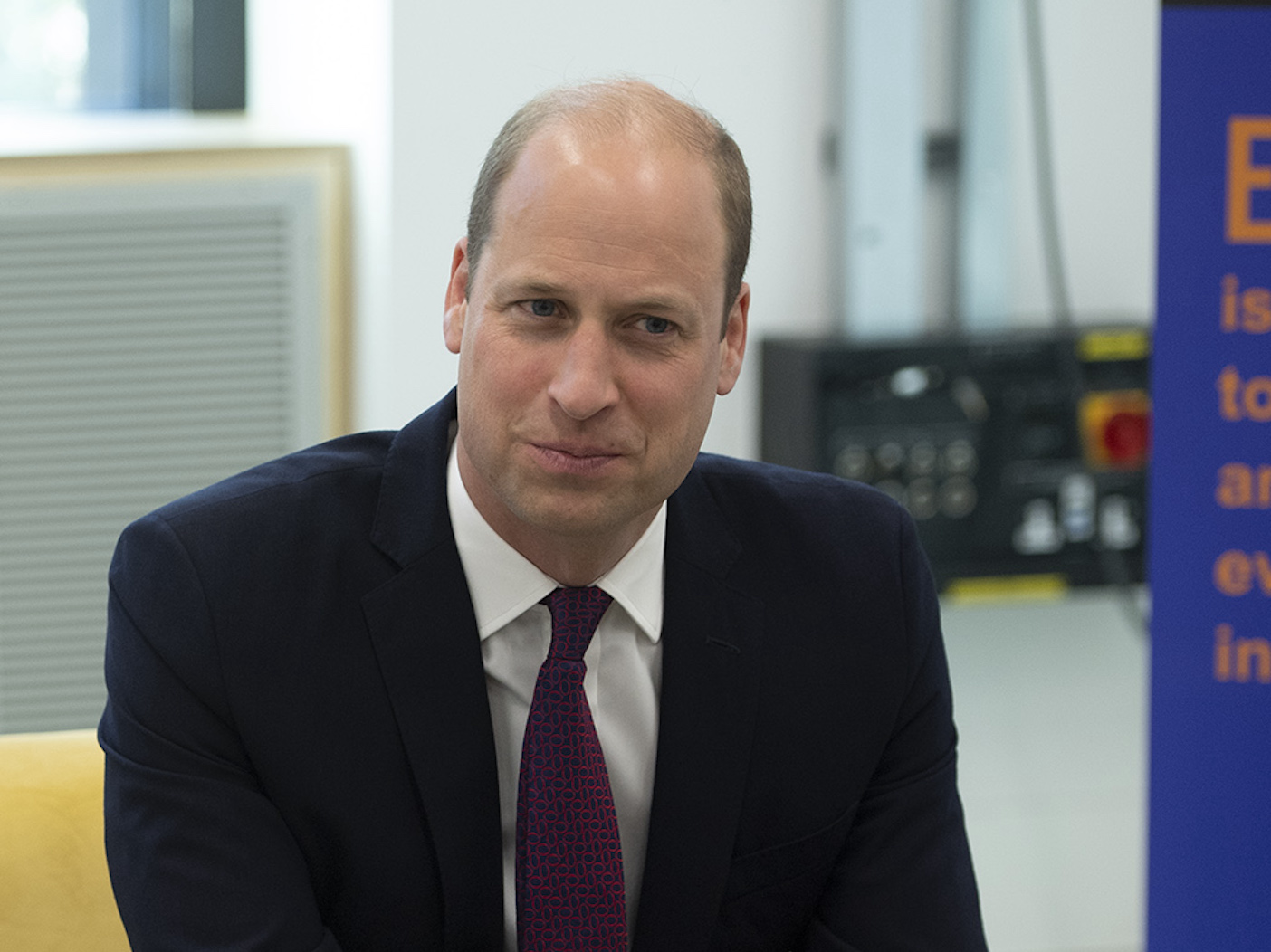 Prince William smiling in a navy suit sitting on a yellow couch