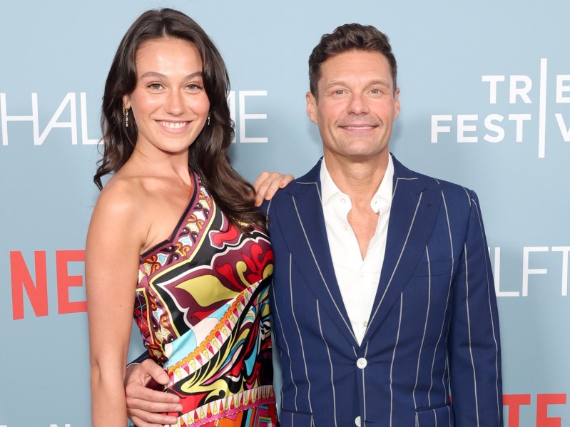 Ryan Seacrest (R) in navy blue suit standing next to Aubrey Paige in multicolored dress against sky blue backdrop
