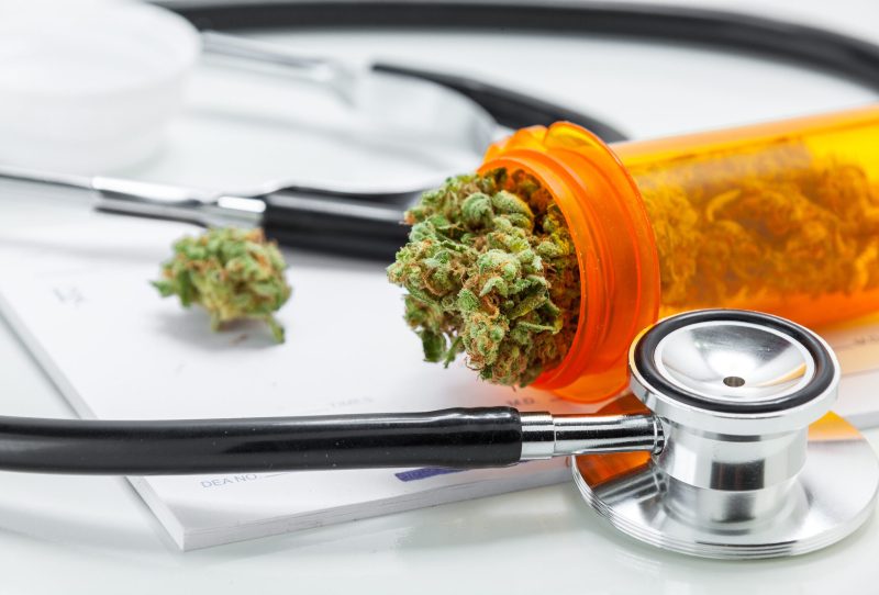 A pill bottle filled with cannabis buds on a doctor's notepad with a stethoscope