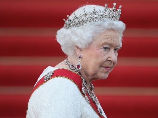 Queen Elizabeth wearing her crown in a white dress in front of a set of red stairs