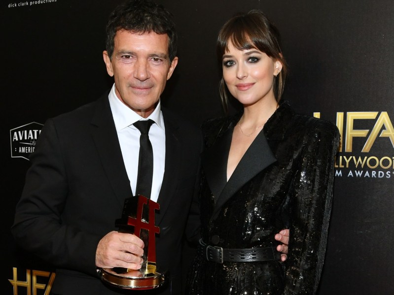 Antonio Banderas (L) in classic black suit and tie, posing with Dakota Johnson, who is wearing a black suit dress