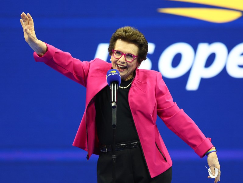 Billie Jean King smiling and waving in a pink jacket at the U.S. Open