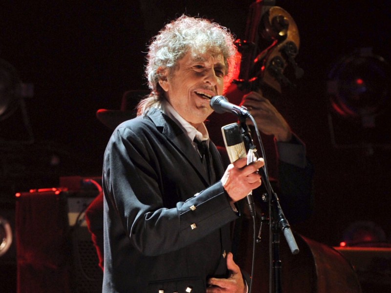 Bob Dylan smiles and sings into microphone onstage in black jacket