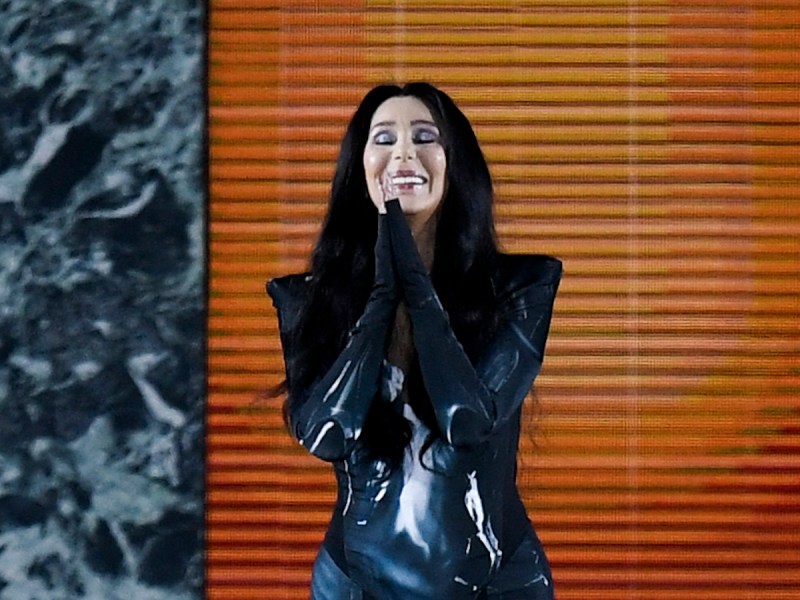 close up of Cher smiling with her hands clasped in front of her in a black outfit on a runway