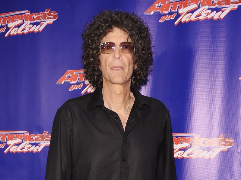 Howard Stern stands in black button-down shirt against blue backdrop