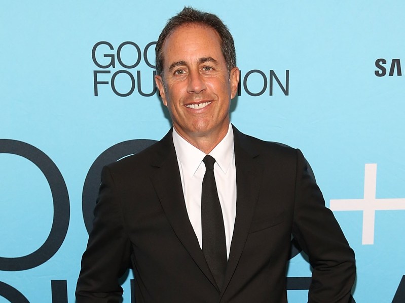 Jerry Seinfeld smiles in classic black suit and tie against light blue backdrop