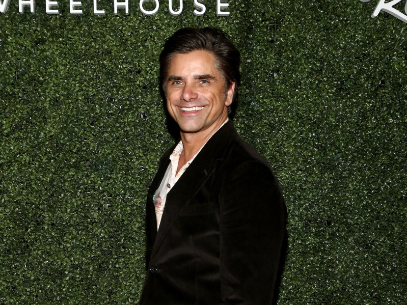 John Stamos smiling in a black suit against a hedge
