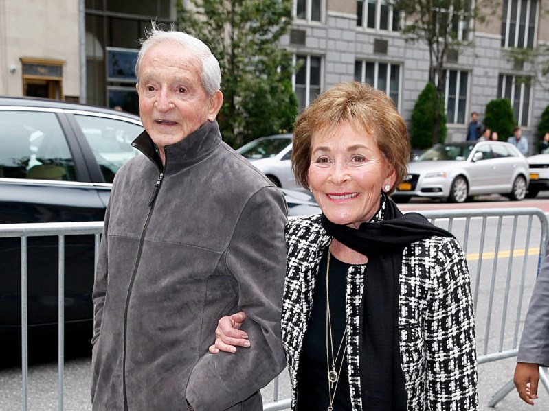 Judge Judy in a black and white sweater smiling arm-in-arm with husband Jerry outside