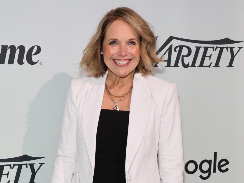 Katie Couric smiles in black top and white blazer against white backdrop