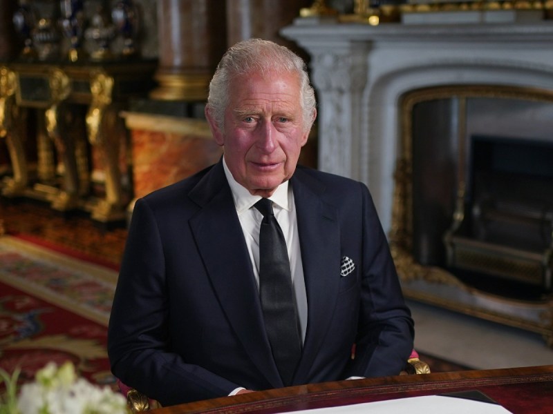 King Charles sits at table in front of fireplace while wearing a navy blue suit and tie