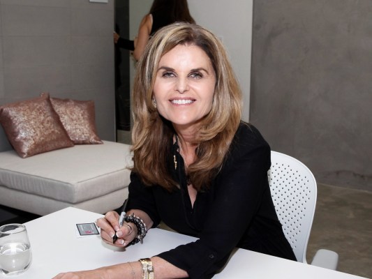 Maria Shriver smiles in black shirt while sitting at table with pen in hand