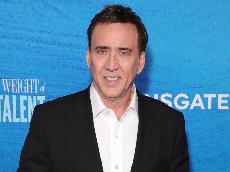Nicolas Cage smiling in black blazer over white dress shirt against blue backdrop