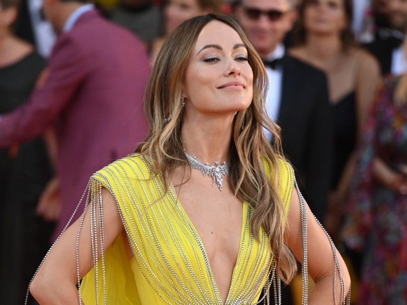 Olivia Wilde smiles with her hands on her hips wearing a yellow gown