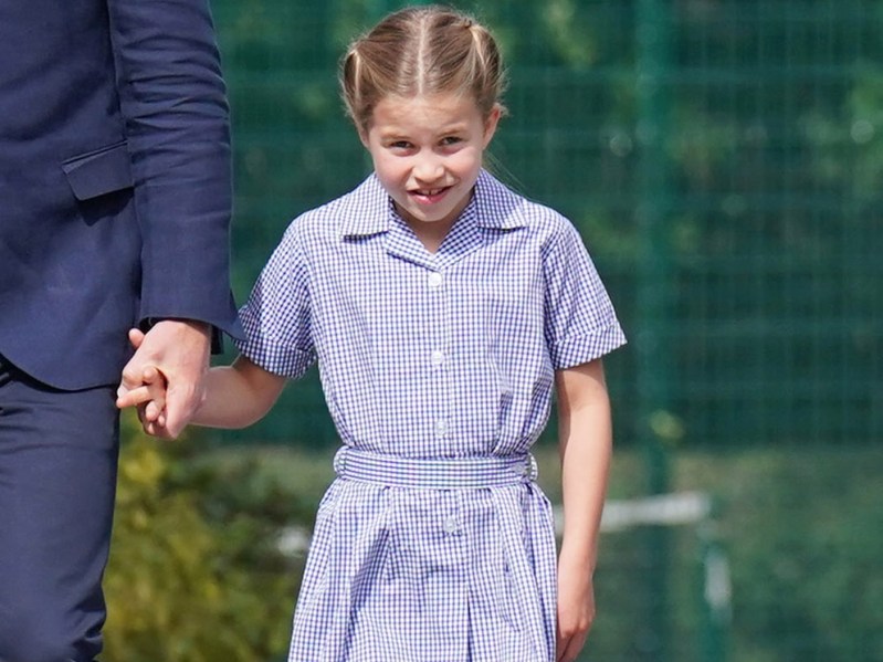 Princess Charlotte walks outside in blue plaid smock dress, holding hands with someone on her right, who is not pictured