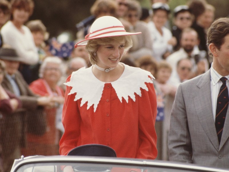Princess Diana smiles in a red top and white hat outside among a crowd