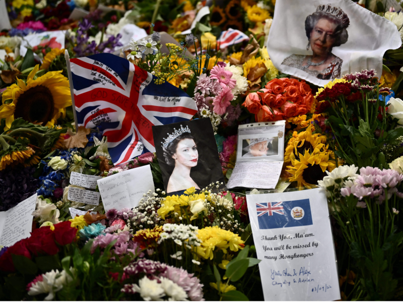 The photo shows a pile of flowers, flags, and photos of Queen Elizabeth that would mourners have left in honor of her passing