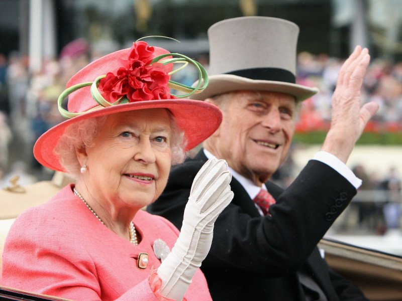 Queen Elizabeth (L) in pink outfit waving alongside Prince Philip, who is in a black suit and gray hat