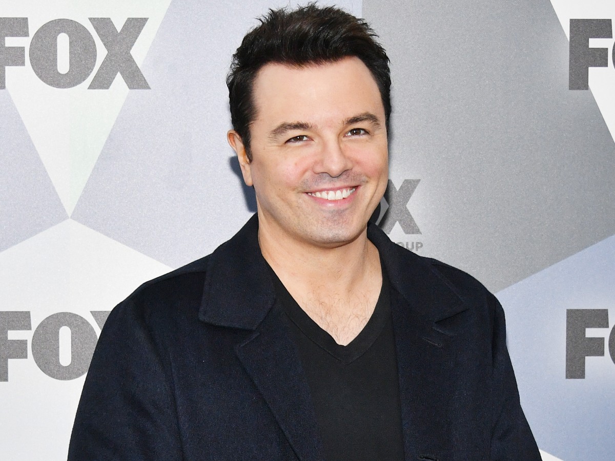 Seth MacFarlane smiles in black top and black jacket against white backdrop