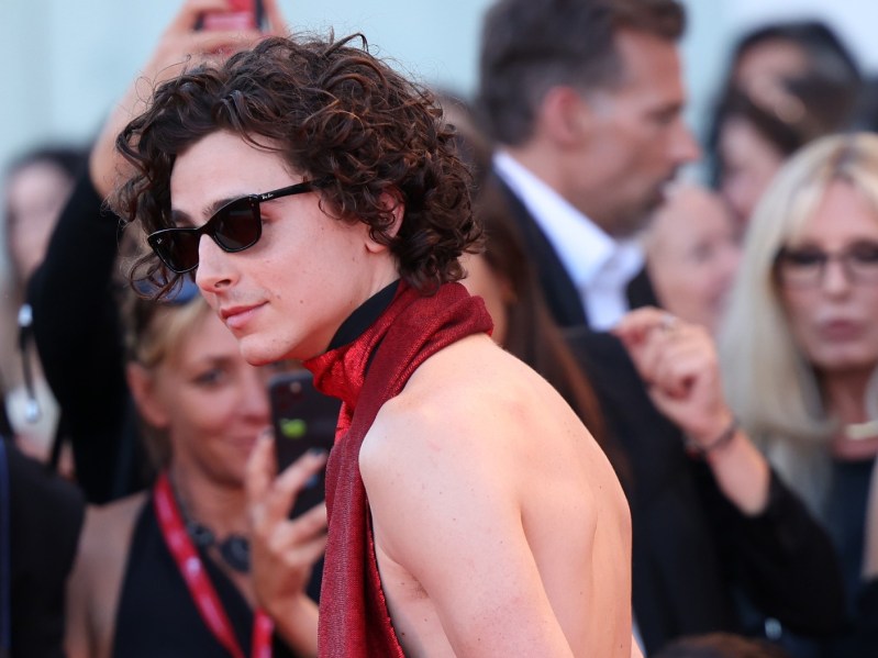Timothée Chalamet standing on red carpet in red backless jumpsuit and sunglasses
