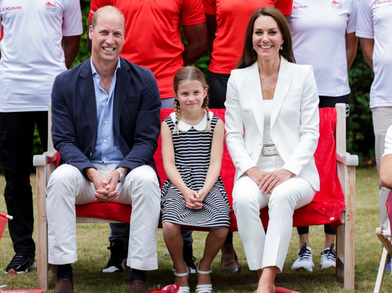 (L-R) Prince William, Princess Charlotte, and Kate Middleton sitting on outdoor bench.