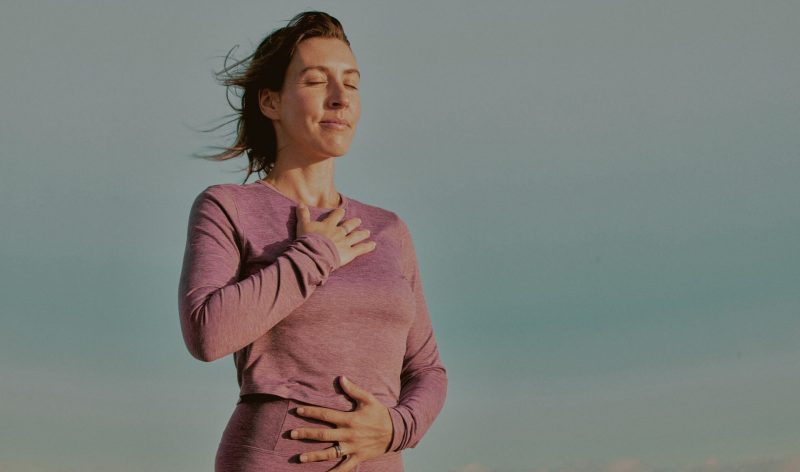 A white woman stands with wind blowing her hair, her hand on her heart, wearing a pink shirt