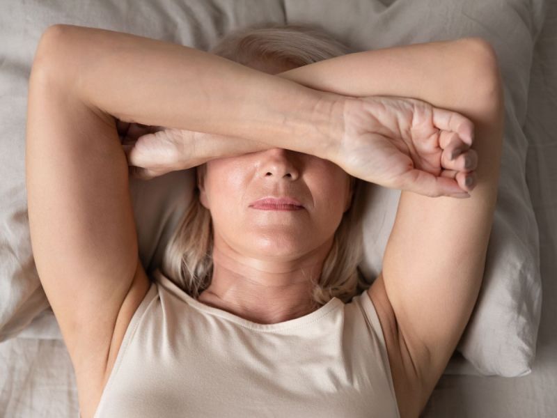 a woman lies on her back in bed with forearms covering her eyes