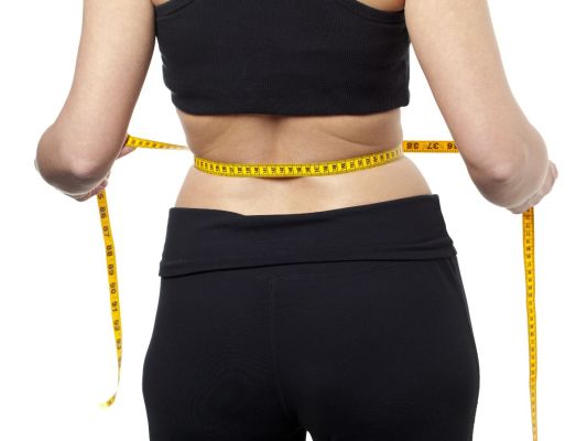 A woman in black workout clothing measures her waist with a tape measure