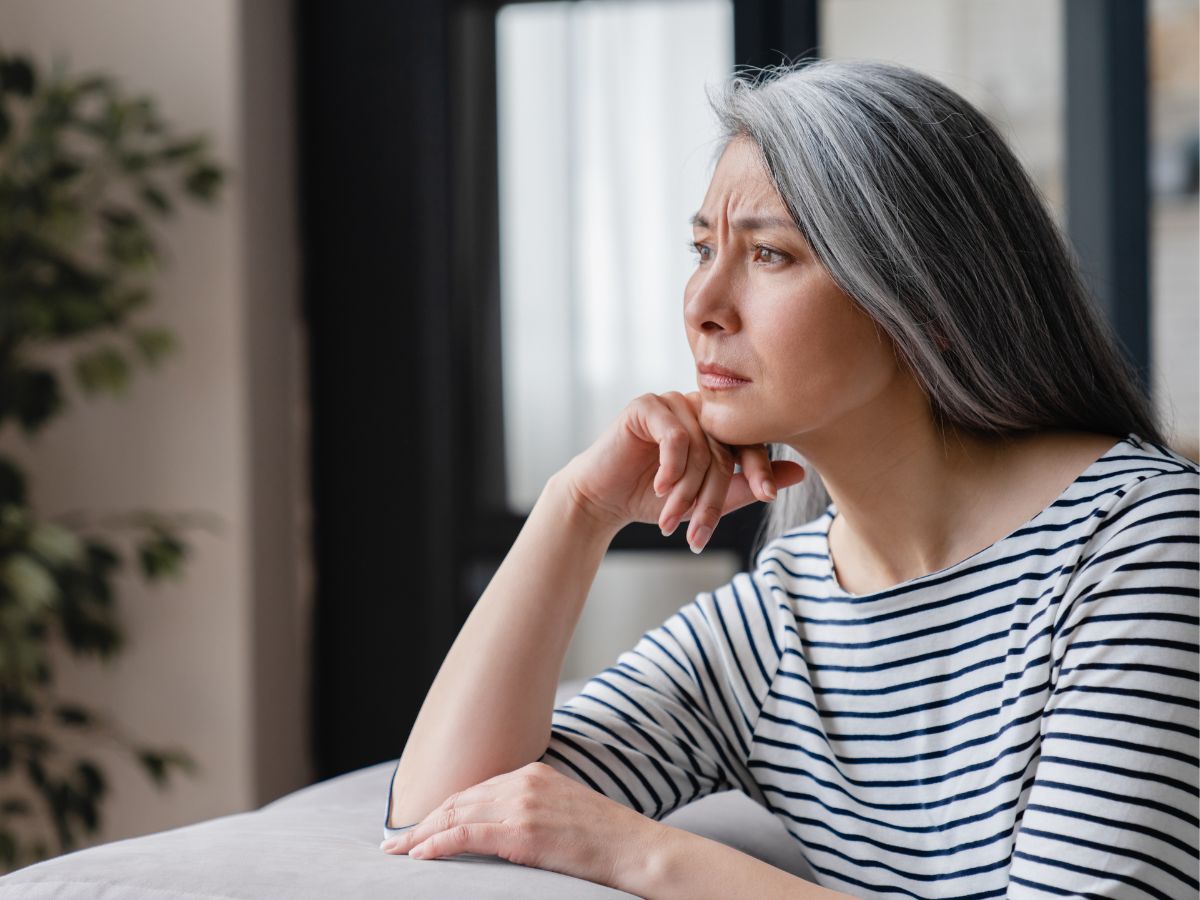 A woman with long gray hair frowns and looks out the window