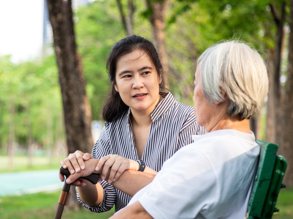 An elderly woman (R) is seen from behind while sitting on a bench. A young woman is facing her and they appear to be in conversation