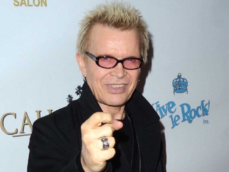 Billy Idol smiles and points at the camera. He is wearing a black jacket and tinted sunglasses