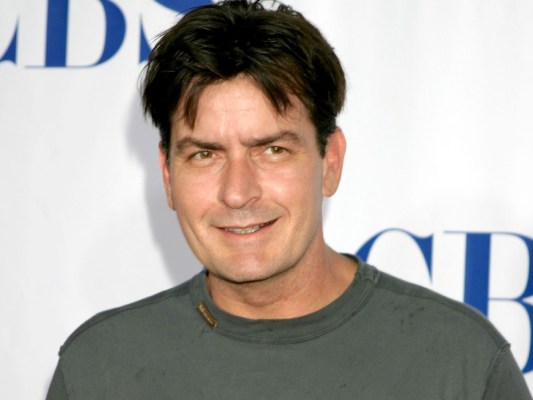Charlie Sheen smiles in olive green tee shirt against white backdrop