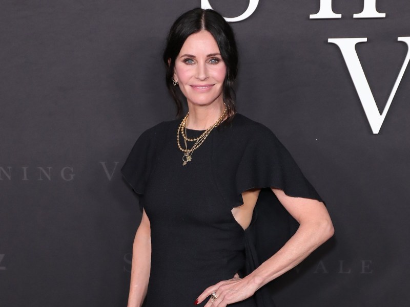 Courteney Cox smiles with her hand on her hip in a black flutter-sleeve dress against a charcoal backdrop