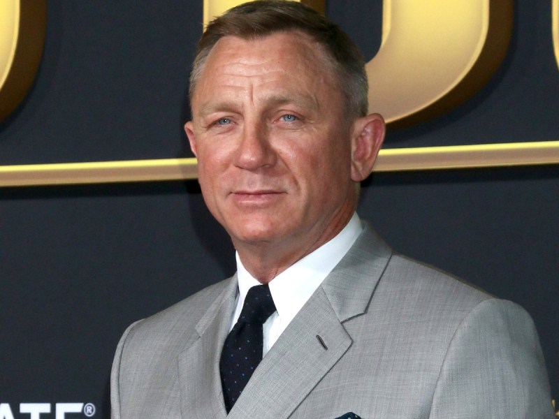 Daniel Craig poses in gray suit with black tie against black/gold backdrop