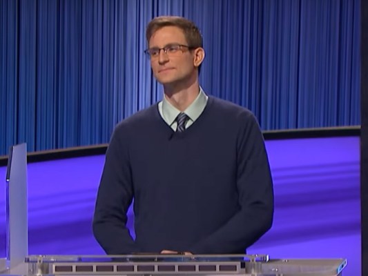 David Sibley stands on "Jeopardy!" stage while wearing dark blue sweater over a dress shirt and tie
