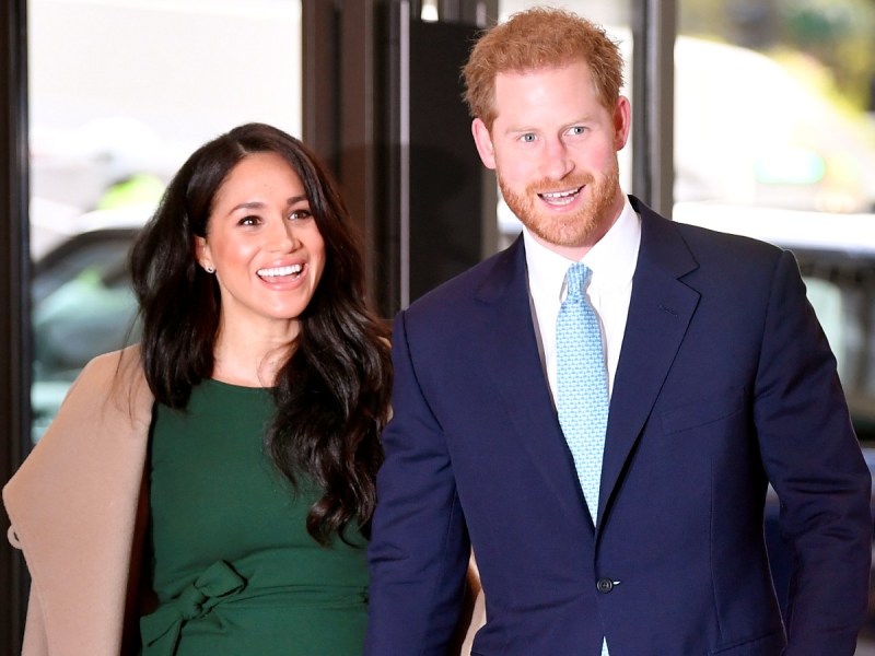 Meghan Markle (L)in green dress with beige coat smiling next to Prince Harry, who is wearing a navy blue suit with light blue tie
